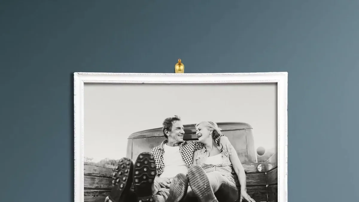 Weiß gerahmtes Schwarzweißfoto eines Paares hängt an einem goldenen FLOREAT-Haken vor blauer Wand